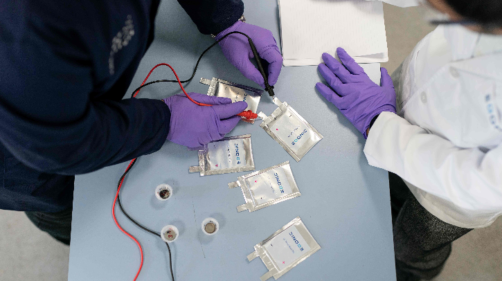 Two scientists in lab coats and purple gloves are working on testing several flat, pouch-like batteries labeled Eqonic with a multimeter