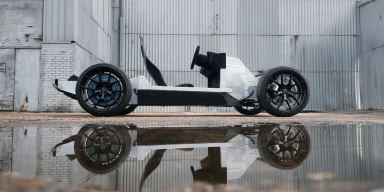 The Watt Electric Vehicle Company PACES electric vehicle skateboard platform shown in prpfile in an industrial settting, with a grey warehouse wall behind, concrete flooring and puddles of water reflecting the vehicle