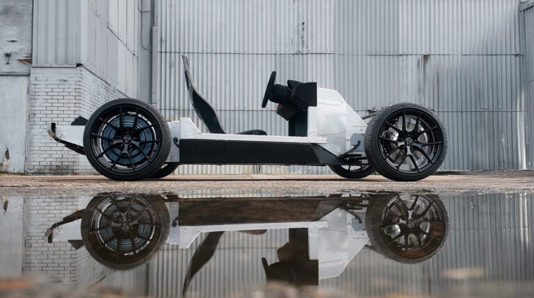 Watt Electric Vehicle Company to integrate Donut Lab’s in-wheel motors into PACES platform The Watt Electric Vehicle Company PACES electric vehicle skateboard platform shown in prpfile in an industrial settting, with a grey warehouse wall behind, concrete flooring and puddles of water reflecting the vehicle