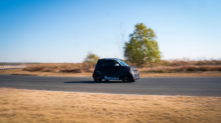 Smart’s all-new electric city car drives along a rural road with short brown grass in the foreground and a solitary tree behind.