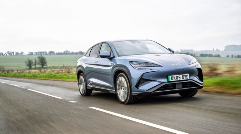 A light blue BYD Sealion drives along a rural road under a grey sky.