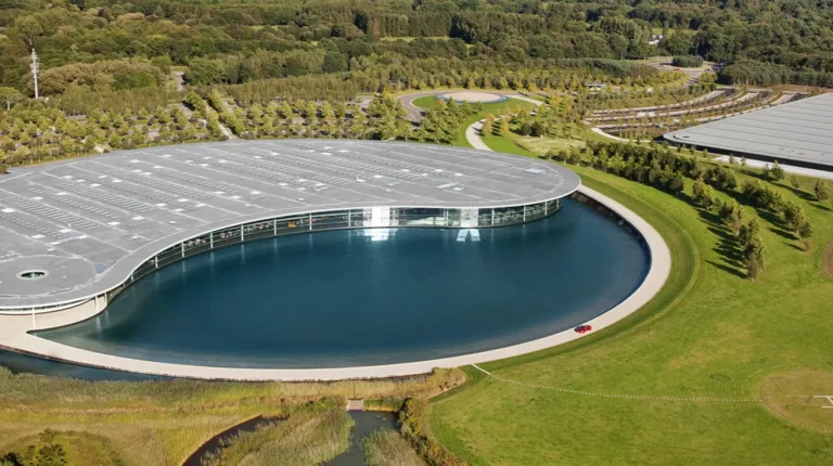 Aerial view of the McLaren Technology Centre in Woking, UK, wiht a large round building and a lake and green grass surrounding these.