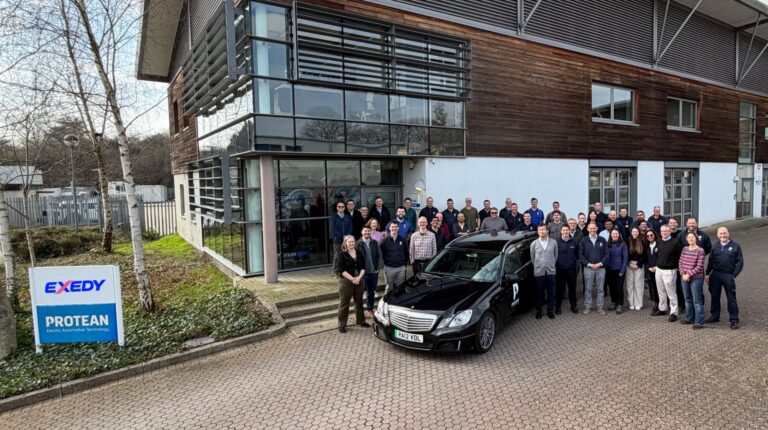 Exedy and Protean staff pose in front of a headquarters building with signage displaying both companies' logos.