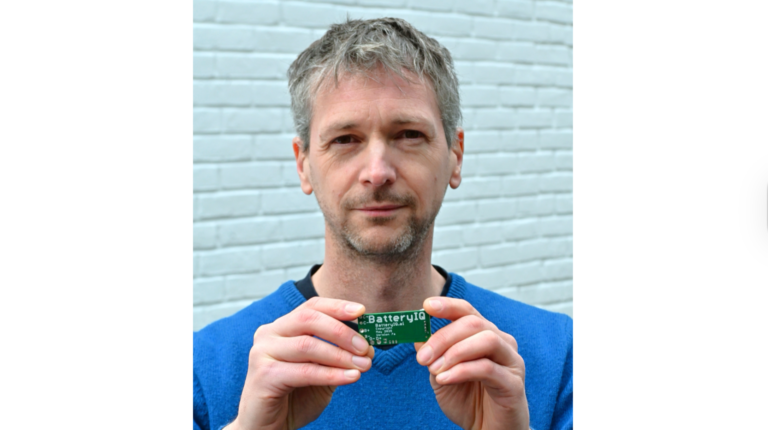 Nick Bailey, founder of BatteryIQ, wearing a blue jumper, standing in front of a light. bue brick wall, and hold a small PCB with the words 'BatteryIQ' on.