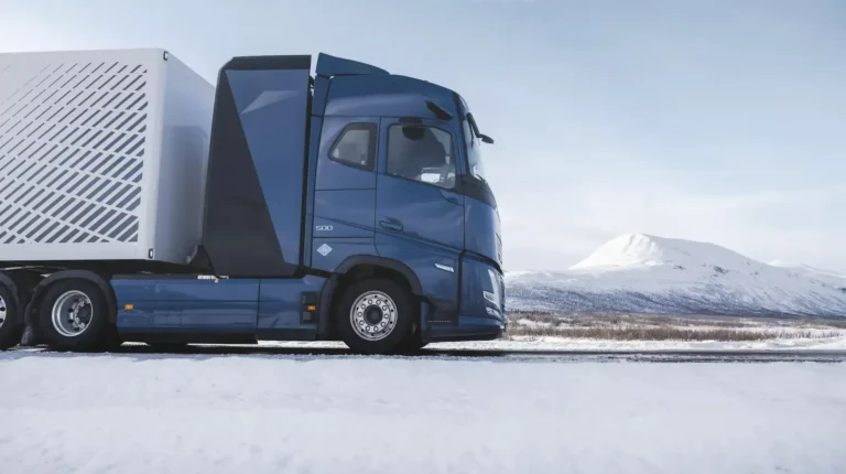 Volvo begins on-road testing of hydrogen ICE heavy trucks A Volvo Trucks hydrogen ICE heavy truck with a dark blue cab and grey trailer undergoes testing in a rural location with snow on the ground and a mountain visible in the background.