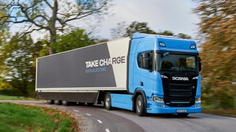 A blue Scania truck pulls a trailer that says "Take Charge - 10% electric". the vehicle is driving along a rural road with a blurred background of trees.