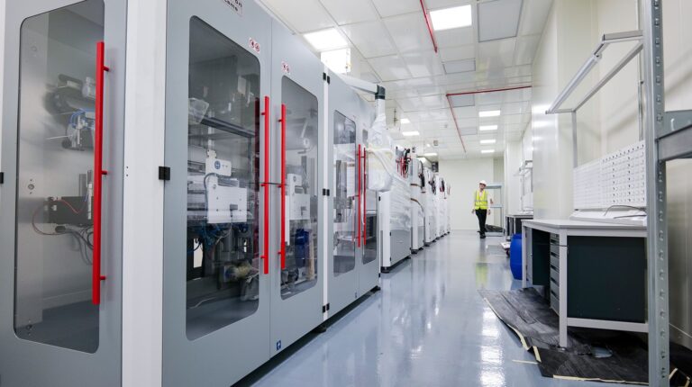 Person in a high-visibility vest stands in a clean, bright laboratory and manufacturing space at the UKBIC, with large industrial machines and empty workbenches.